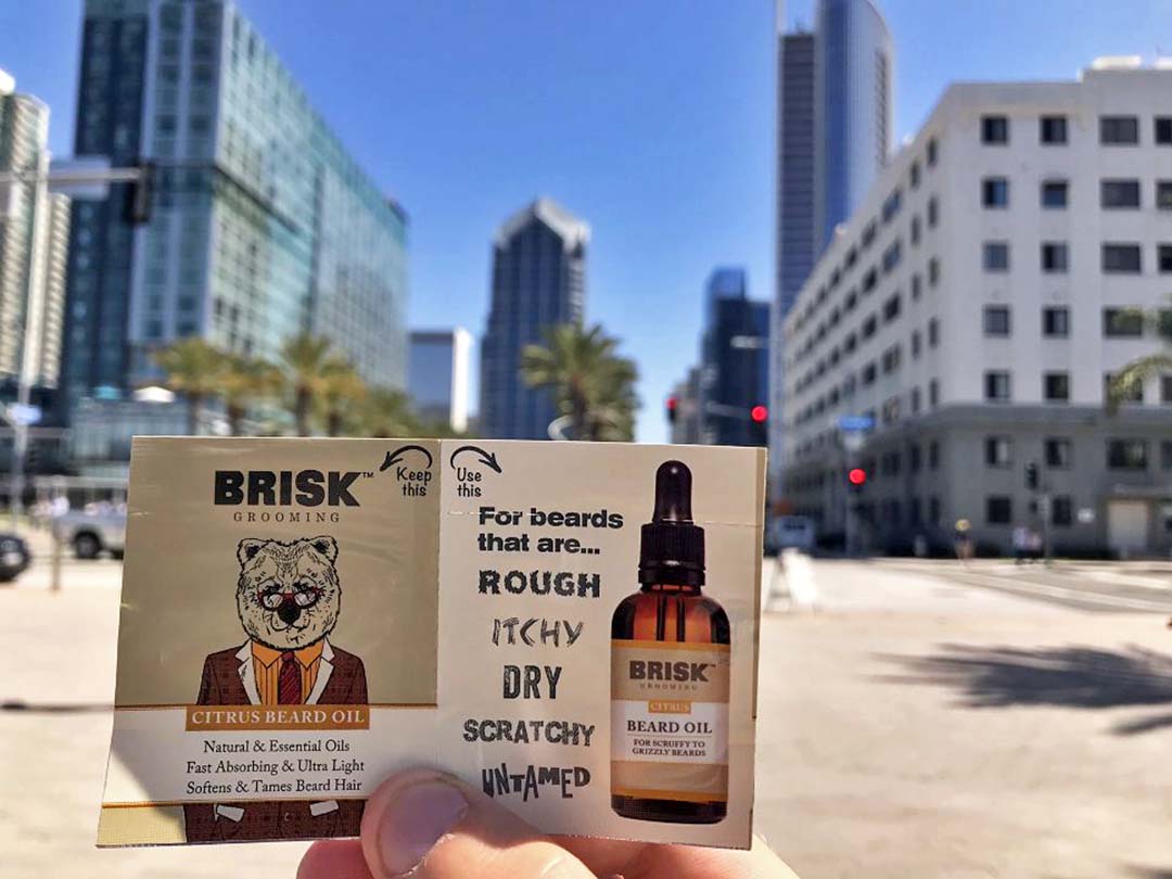 Close up of a person’s hand holding a Brisk beard oil sample with cityscape in the background